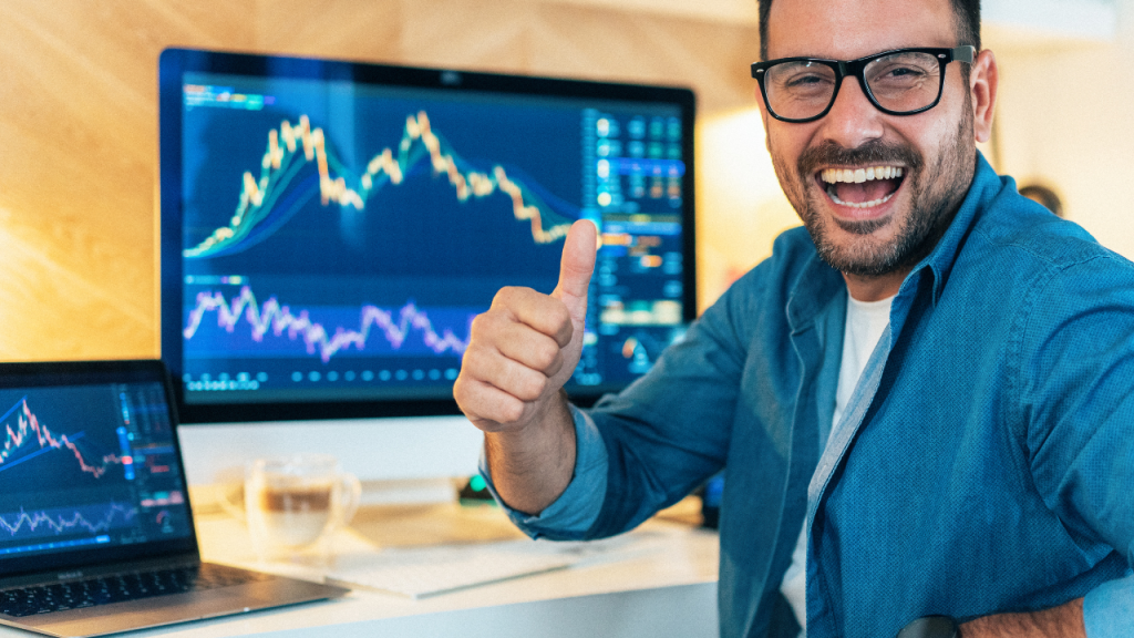 モニターとノートパソコンで株価を見てこちらを向きながら笑っている男性の画像