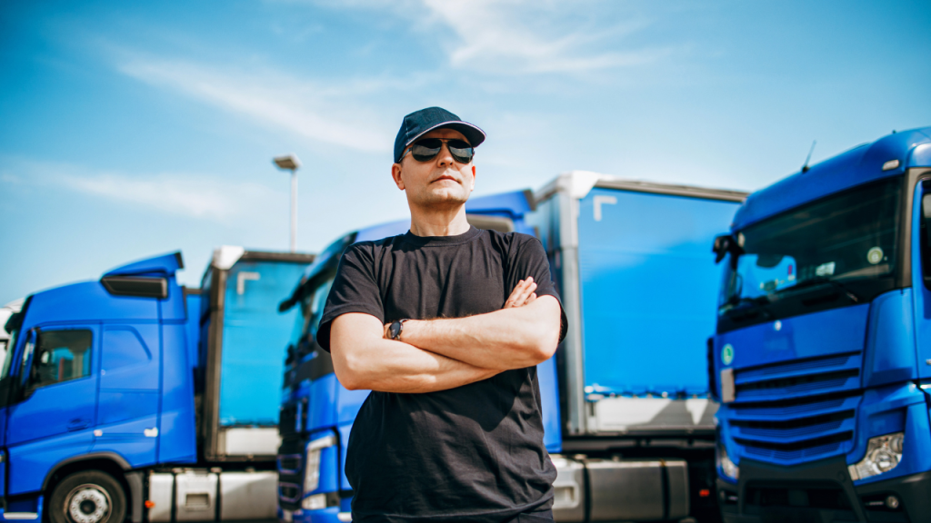 青空の元、青いとのトラックが3台駐車されている。その正面に男性がサングラスをかけ腕組みしている画像。